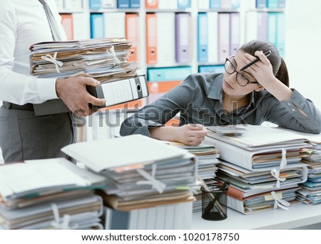 Young stressed secretary in the office overwhelmed by work and desk full of files, her boss is bringing more paperwork to her Royalty-Free Stock Photo #1020178750