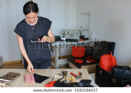 Young woman using mobile to take pictures of handmade notebook with black cover. Married couple do business connected with making leather bags backpacks belts wallets. Female wearing grey T-shirt