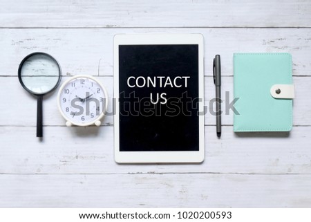 Top view of magnifying glass,table clock,pen,notebook and tablet computer written with ' CONTACT US' on white wooden background.