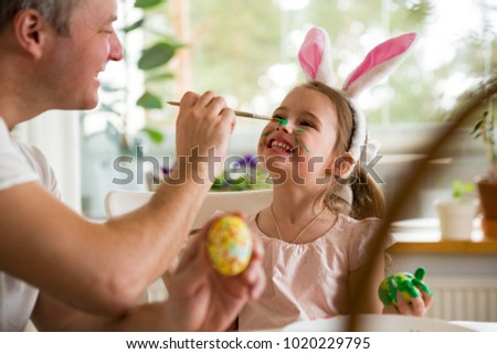 A father and daughter celebrating Easter, painting eggs with brush. Happy family smiling and laughing, drawing on face. Cute little girl in bunny ears preparing the holiday. Royalty-Free Stock Photo #1020229795