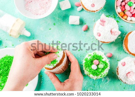 Making easter cupcakes, person decorate cakes with bunny ears and candy eggs, copy space frame top view, girl's hands in picture