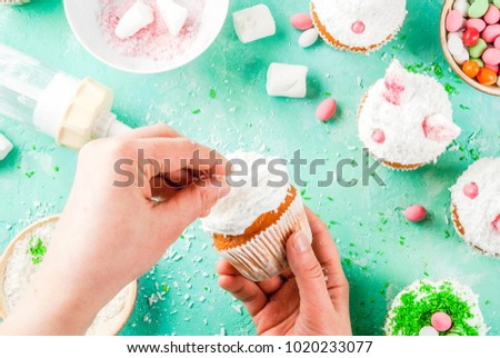 Making easter cupcakes, person decorate cakes with bunny ears and candy eggs, copy space frame top view, girl's hands in picture