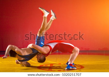 Two  strong men in blue and red wrestling tights are wrestlng and making a suplex wrestling on a yellow wrestling carpet in the gym. Wrestlers doing grapple. 