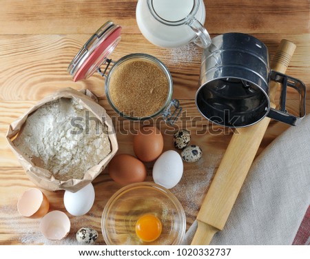 Set for baking. In the picture, a jug of milk, flour, quail eggs and eggs, a rolling pin, a flour sifter, a yolk with protein in a bowl, and an egg shell. Wooden background. Close-up. View from above.