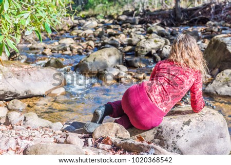 Woman back in red lying down taking pictures of shallow rock stream with running water and waterfall in autumn with green leaf foliage, stones and flowing creek