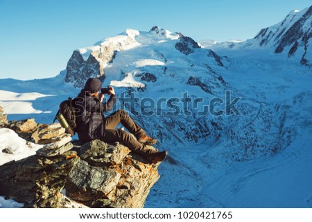 Traveler Man with backpack trekking in mountains, enjoy beautiful view and making photo on smartphone. Explorer man hiking, travel in Alps hills, Switzerland. Hiker sitting on rock cliff outdoors.