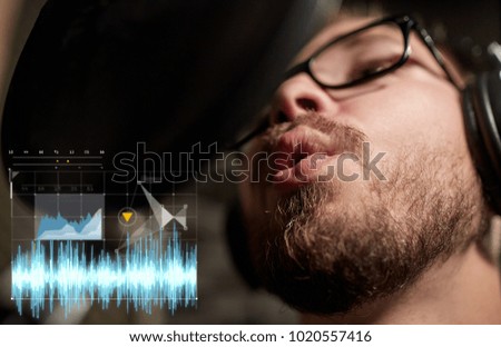 music, show business, people and technology concept - male singer with headphones and microphone singing song at sound recording studio