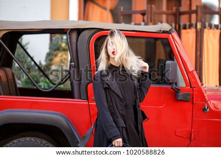 Street fashion concept. Trendy look of stylish young blond woman in black fashion coat and hat. Young lady standing over red car. Sunny spring or autumn weather. Glamour red lips