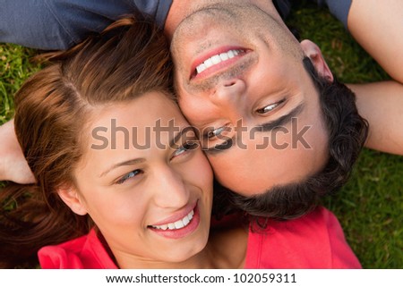 Close-up of two friends smiling while looking at each other as they lie head to shoulder with an arm behind their head on the grass