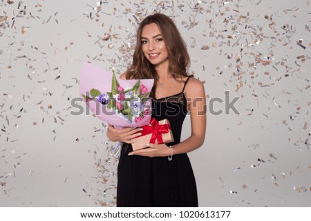 Charming, beautiful girl in black dress is  holding cute tulip bouquet. Lady has a tiny box with a present. Confetti.