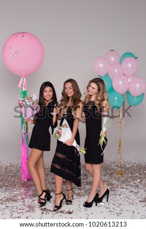 Three beautiful girls in black dresses are holding tulip bouquets. Ladies are drinking champagne. Colorful balloons. Confetti.