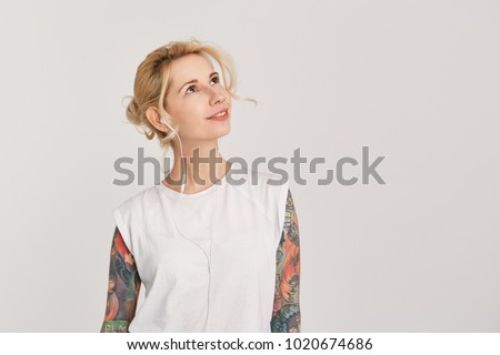 Horizontal portrait of blond, european girl with pierced nose, blue eyes, tattoo on arms, dressed in casual white t-shirt, listen music via headphones and looking aside and above. Copyspace. Isolated