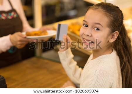 Close up of a cheerful cute little Asian girl paying with a credit card at the bakery store smiling to the camera over her shoulder copyspace payment banking money consumerism customer buying