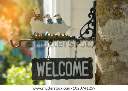 Wooden welcome sign hanging under three duck statues in tree background