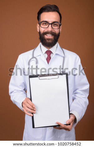 Studio shot of young handsome bearded Persian man doctor against brown background