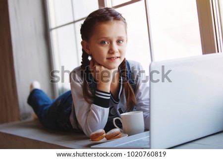 Young cute emotional teenager girl with braids in overalls lies by the window indoor. With cup and cookies, working with the laptop