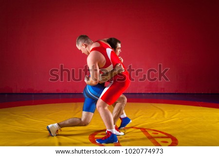 Two strong wrestlers in blue and red wrestling tights are wrestlng and doing grapple on a yellow wrestling carpet in the gym. 