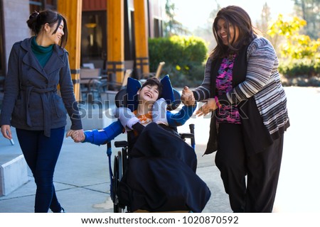 Eleven year old biracial disabled boy in wheelchair holding hands with caregivers while on a walk outdoors Royalty-Free Stock Photo #1020870592