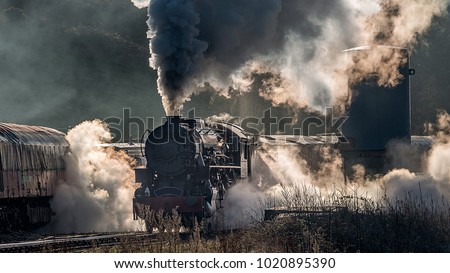 Steam train locomotive approaching a station passing through a goods yard letting of smoke and steam lit from behind creating atmospheric photograph Royalty-Free Stock Photo #1020895390
