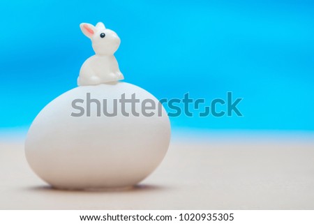Little white toy rabbit sitting on a big white egg on a blue background