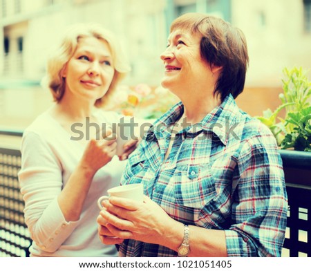 Smiling elderly female friends staying on balcony with cup of coffee
