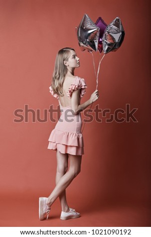 Full length portrait of young woman wearing pink dress with opened back holding silver balloons in studio on red background. Celebrating concept.
