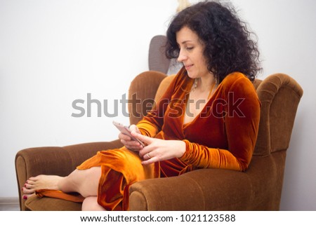A 40-year-old woman sits in a golden velvet dress sits in a soft brown chair with a phone in her hands