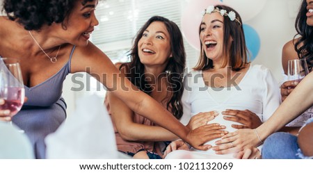 Excited young female friends touching pregnant woman's belly at baby shower party. Laughing pregnant woman sitting with friends touching her tummy. Royalty-Free Stock Photo #1021132069