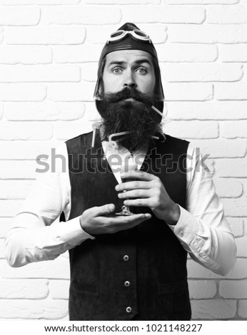 handsome bearded aviator man with long beard and mustache on surprised face holding glass of alcoholic shot in vintage suede leather waistcoat with hat and glasses on white brick wall background