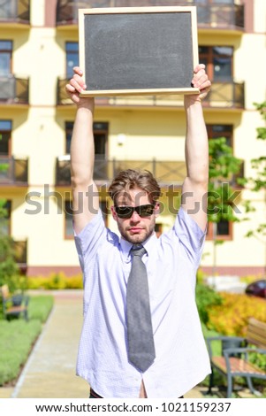 Salesman with advertise for sale. Business and real estate concept. Estate agent holds blackboard or chalkboard above his head, copy space. Businessman with calm face with building on background.