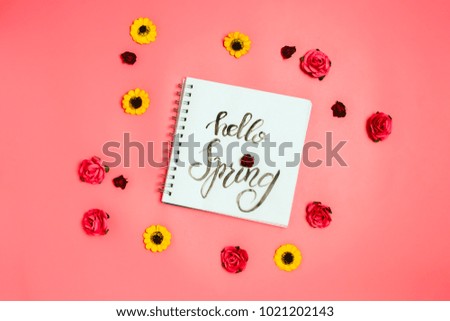 Notebook with white page with handlettering "Hello Spring" and various artificial flowers around. Flatlay,  pink background. 