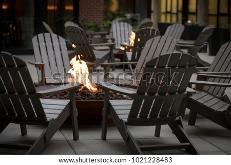 Low angle view of a fire pit surrounded by a circle of Adirondack chairs, with another campfire and hotel building in the dark background Royalty-Free Stock Photo #1021228483
