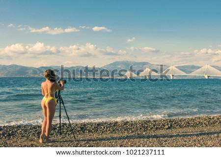 Young female in the bikini on the beach with tripod and camera taking picture of Rion-Antirion bridge near Patras, Greece
