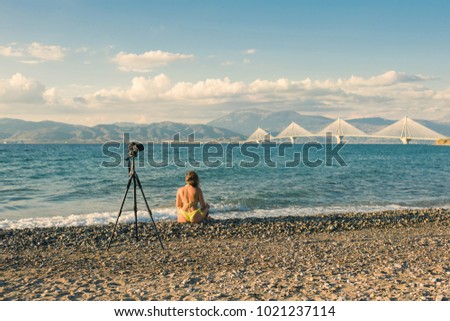 Young female in the bikini on the beach with tripod and camera taking picture of Rion-Antirion bridge near Patras, Greece