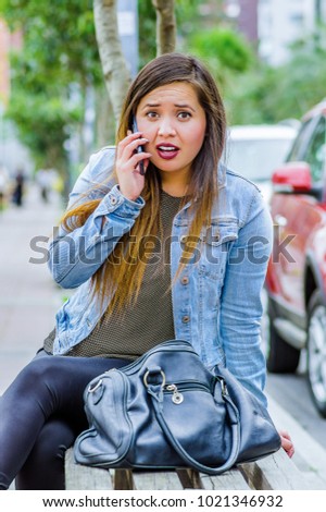 Fashion trendy casual young woman wearing a jean jacket and black leggings, using her cellphone and showing shock emotion. Female portrait with shocked facial expression