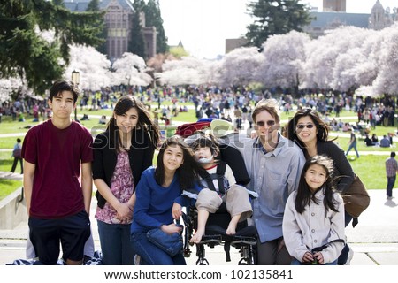 Multiracial family of seven in front of crowded field of cherry blossom trees. Youngest child is disabled with cerebral palsy.