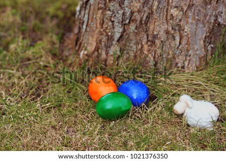 three Easter eggs and a white ceramic sheep in the grass beside a tree trunk, close-up