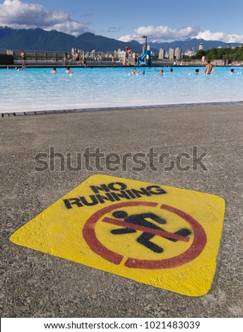 A bright yellow No Running sign painted on concrete is in the foreground, with a blue swimming pool and Vancouver's North Shore Mountains in the background.
