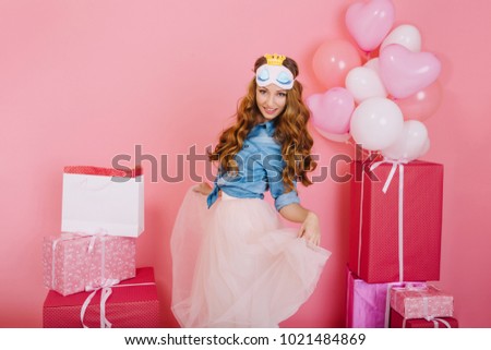 Portrait of elegant girl in sleep mask celebrating her birthday and dancing next to present boxes. Charming long-haired young woman received many gifts and enjoys event with balloons on background