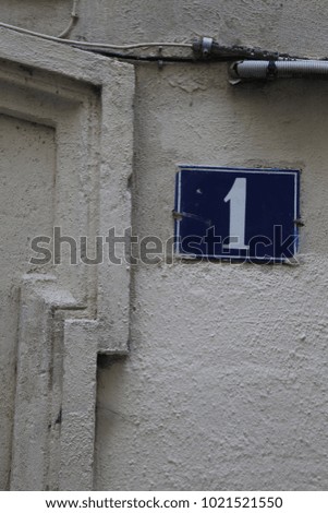 Close up view of the number one written on a blue rectangle fixed on the wall of an old house. Decorative elements and lines on the beige painted wall. Numeric white symbol in a city street in France 