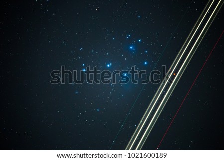 a plane flies in the night sky, in the background the stars of the Pleiades light up