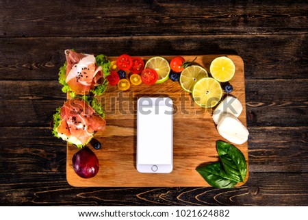 Traditional Italian food named prosciutto, lies on the cutting board, along with biologically grown vegetables. With blank screen on smart phone.