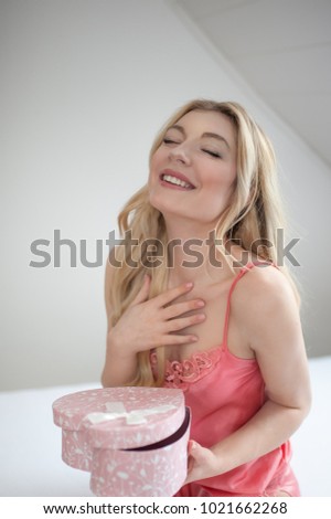 Beautifil young woman with blonde long hair is lying in her white bed holding pink rose and looking into the gift box in the shape of heart. She is happy about the gifts to Valentines Day or Birthday