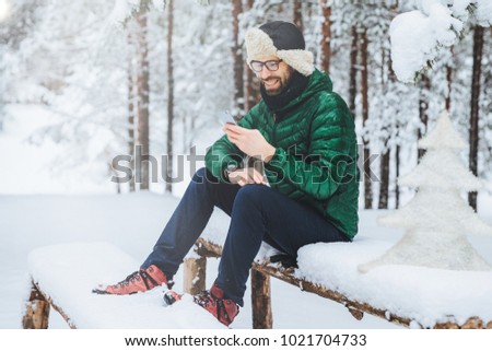 Smiling positive male wears warm winter clothes, reads message on mobile phone, spends free time in calm atmosphere outdoors in winter forest, enjoys fresh air. People, rest, technology concept