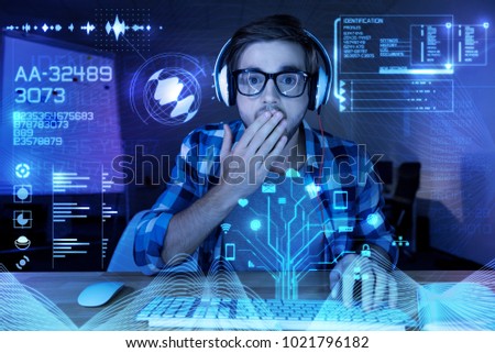 Oh no. Emotional young skilled programmer closing his mouth with his hand and feeling surprised while looking at the modern transparent screen and reading unpleasant information about cyber security