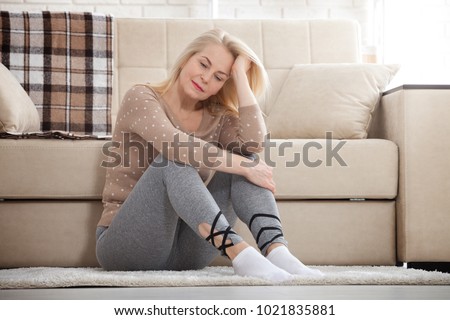 Depression. Middle aged barefoot woman sitting at the floor embracing her knees, near sofa at home, her head down, bored, troubled with domestic violence.