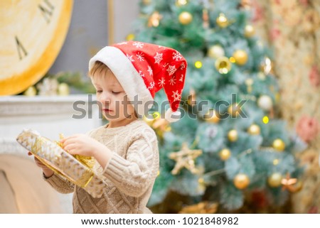 a little boy in a red cap sits near a Christmas tree with a gift