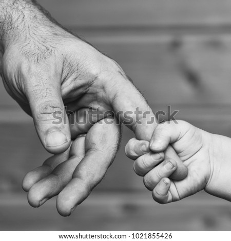 Closeup of two touching hands of small baby boy holding finger of male father as symbol of family love and trust on blurred wooden background, square picture