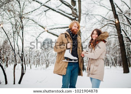 Winter and date. Young couple, lovers man and woman in winter on a background of snow and forest holding hands and smiling. A guy with long hair and a beard is holding skates and looking at the girl.