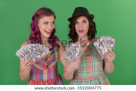 Two emotional and beautiful young girls in bright summer dresses, holding in their hands dollar bills won in a lottery against the background of a green wall.The girl steals the dollar